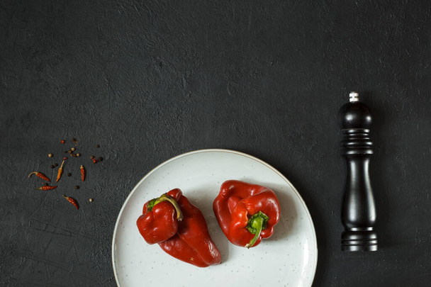 Δύο άσχημες κόκκινες πιπεριές κατάλληλες για μαγείρεμα σε γκρι πιάτο. Μαύρο φόντο με μύλο πιπεριού και μπαχαρικά, κορυφαία άποψη με χώρο - Φωτογραφία, εικόνα