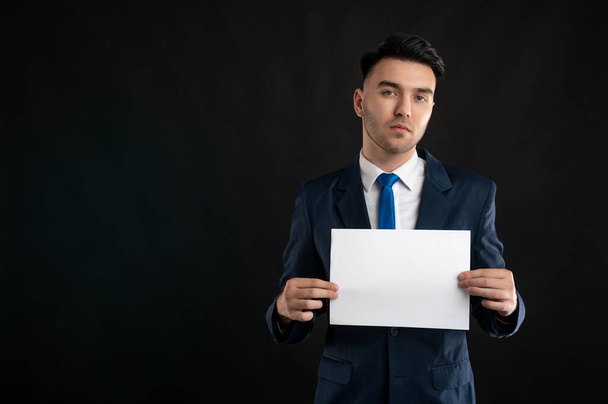 Retrato del hombre de negocios con traje de negocios azul y corbata sosteniendo papel blanco aislado sobre fondo negro con área de publicidad de espacio de copia - Foto, Imagen