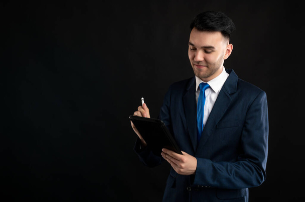 Portret biznesmena w niebieskim garniturze i krawacie, notatki na tablecie odizolowanym na czarnym tle z przestrzenią reklamową. - Zdjęcie, obraz