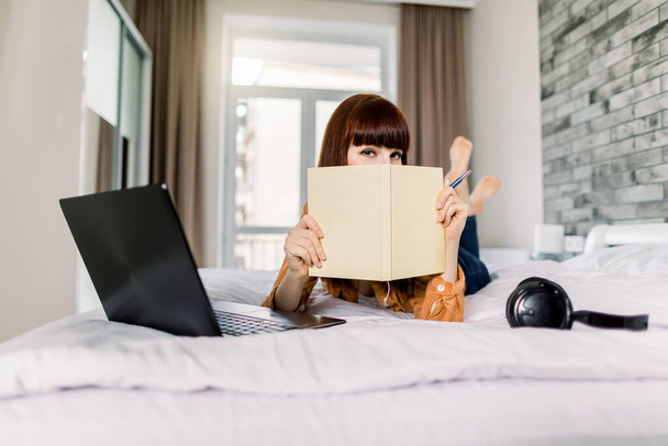 Young beautiful woman in casual wear, hiding her face with notepad or book, while working on a laptop, lying on the bed at home. Headphones and laptop on bed. Freelance, work from home concept - Φωτογραφία, εικόνα