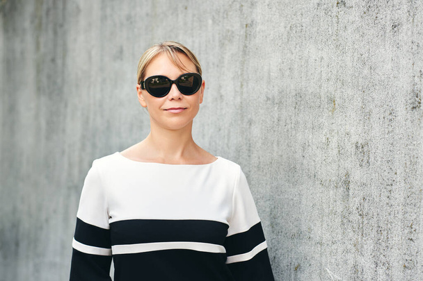 Outdoor portrait of pretty middle age woman wearing black and white dress and sunglasses, posing on grey wall background - Photo, Image