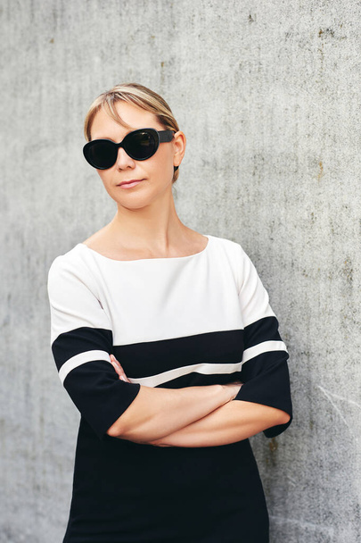 Внешний портрет красивой женщины средних лет в черно-белом платье и солнечных очках, позирующих на сером фоне стены - Фото, изображение