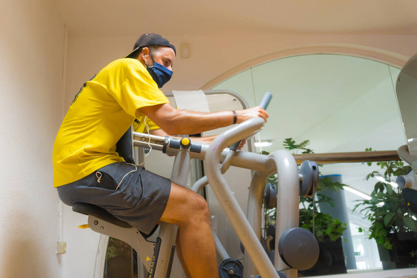 Caucasian young man with face mask on a gym machine performing exercises, gym with little vacuum, new normal, social distance, covi-19, coronavirus, pandemic - Фото, изображение