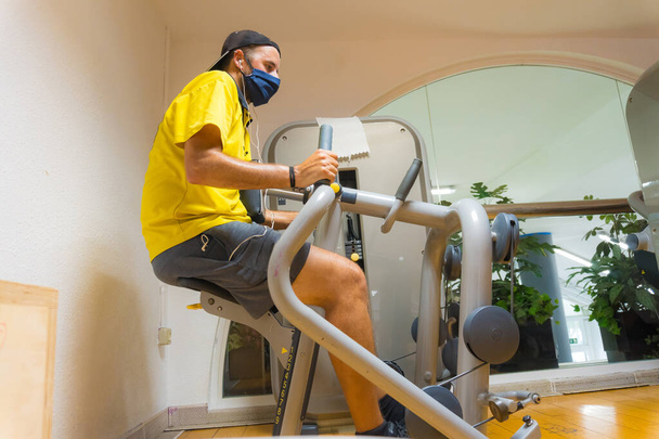 Caucasian young man with face mask on a gym machine performing exercises, gym with little vacuum, new normal, social distance, covi-19, coronavirus, pandemic - Fotoğraf, Görsel