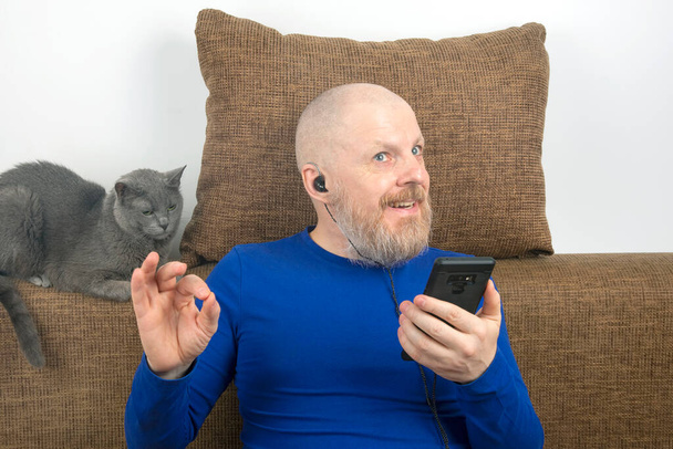 γενειοφόρος άνθρωπος ακούει με ευχαρίστηση στο σπίτι την αγαπημένη του μουσική από το τηλέφωνο με μικρά ακουστικά. Γκρίζα γάτα. ακουστόφιλος και λάτρης της μουσικής. μουσική και hi-fi ήχος - Φωτογραφία, εικόνα