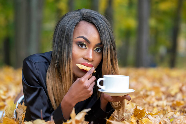 Beautiful young African girl eating cookies and drinking coffee from white Cup on autumn leaves background in Park - Photo, Image
