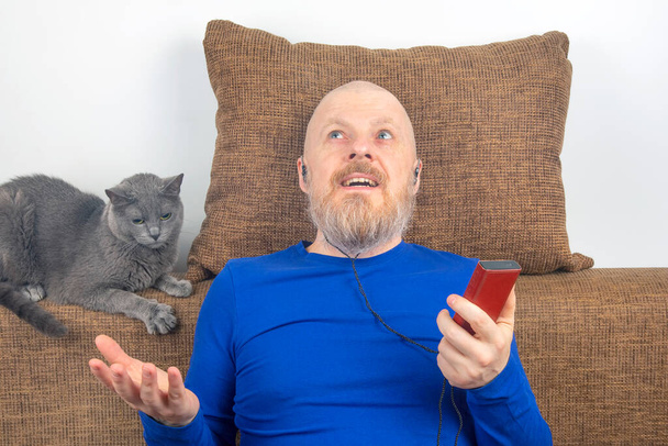 bearded man listens with pleasure at home to his favorite music from the phone with small headphones. gray cat. audiophile and music lover. music and hi-fi sound. - Photo, Image