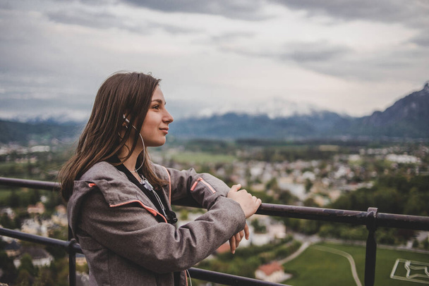 Καυκάσιος μελαχρινή κοπέλα ακούσετε μουσική σε ακουστικά στο παρασκήνιο της πόλης Σάλτσμπουργκ & Άλπεις βουνά με ουρανό σύννεφο. Αυστρία - Φωτογραφία, εικόνα