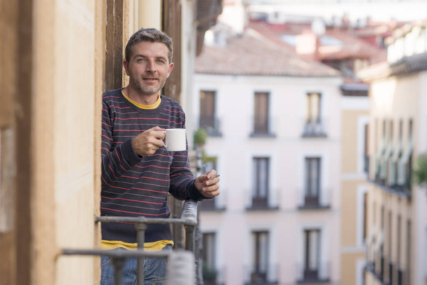 30s or 40s attractive and happy man at home balcony relaxed and cheerful enjoying cup of coffee smoking cigarette looking to city street smiling and enjoying the urban view - Foto, immagini