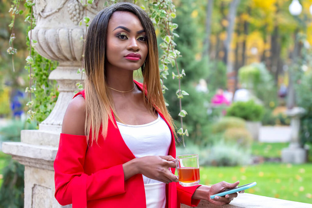 Όμορφη νεαρή Αφρικανή με ένα φλιτζάνι τσάι και ένα τηλέφωνο στα χέρια της στο πάρκο. - Φωτογραφία, εικόνα