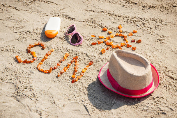 Inscription and shape of sun made of amber stones, sunglasses, sun lotion and straw hat on sand at beach. Sunbathing, summer and vacation time - Photo, image