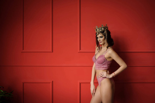close up photo of a sexy fashion woman in pink underwear with crown on her head on a red background in a photo studio - Photo, Image