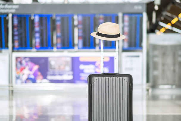 torba bagażowa z kapeluszem w międzynarodowym terminalu lotniskowym, walizka na wózek z tablicą informacyjną w tle lotniska. Pojęcia transportu, podróży i wakacji - Zdjęcie, obraz