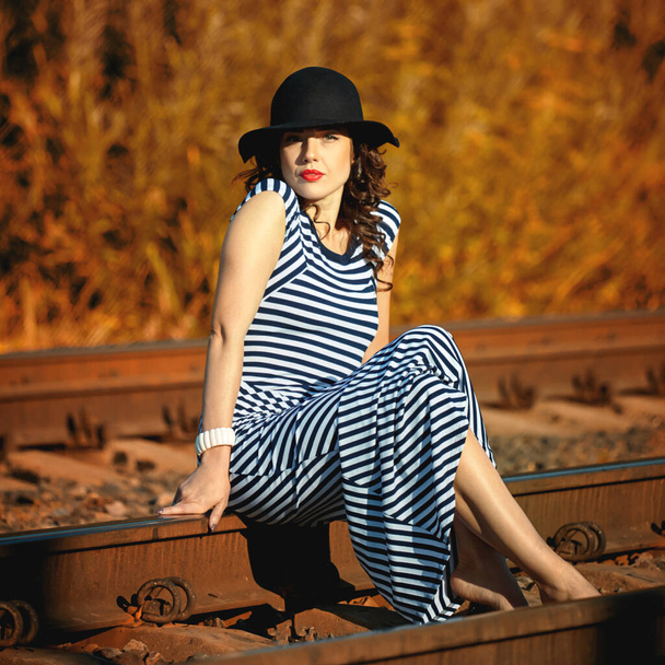 attractive girl in a striped dress and hat sits on the railroad - Zdjęcie, obraz