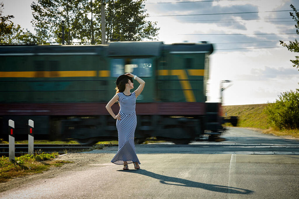 attractive girl in a striped dress and hat posing against the background of a passing train - Fotoğraf, Görsel