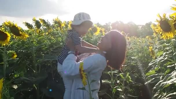 Eine fröhliche Mutter hält ihren kleinen Sohn in den Händen und lacht im Sonnenblumenfeld. Attraktive Frau mit einem kleinen Jungen im Arm auf einem Feld mit Sonnenblumen. - Filmmaterial, Video