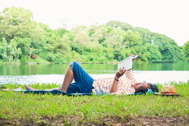 Una anciana yacía leyendo un libro en un parque con un gran estanque. Una vida feliz y sencilla en la jubilación. Concepto comunitario senior - Foto, imagen