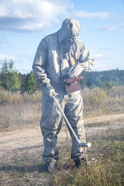Солдат в защитном костюме проверяет радиоактивную территорию дозиметром на фоне дикой растительности и неба с облаками. Дым от вредных испарений распространяется. - Фото, изображение