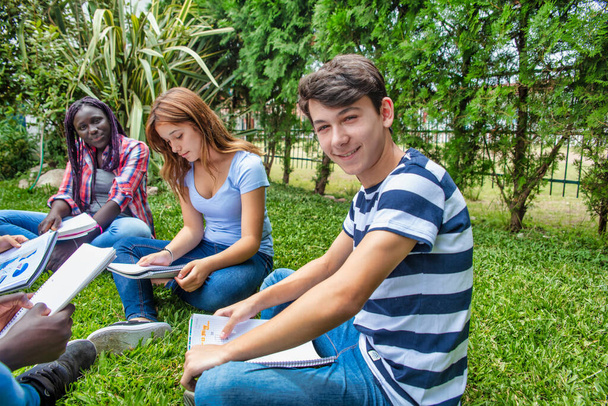 草の上に座って学校のレッスンを作る混合レースのティーンエイジャーの幸せなグループ。学校のコンセプトに戻る - 写真・画像