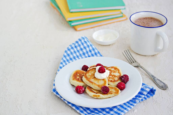 Breakfast, pancakes with sour cream and raspberries on a white plate on a light concrete background. Child's breakfast in front of the school. School breakfast concept. - Φωτογραφία, εικόνα