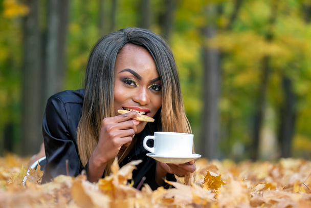 Mooi jong Afrikaans meisje eten koekjes en het drinken van koffie uit witte Beker op herfst bladeren achtergrond in Park - Foto, afbeelding