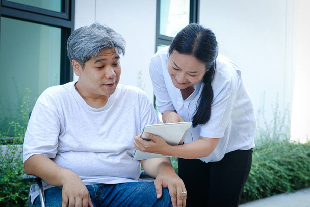 Ein älterer Patient, der im Rollstuhl saß, wurde von einer Krankenschwester betreut. Schauen Sie auf das lächelnde Tablet, lachen Sie, wählen Sie ein Gesundheitscheck-Programm. - Foto, Bild