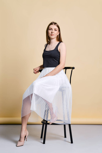 Pretty young woman in black tank top and white skirt sitting on tall chair over pale yellow background - Foto, Bild
