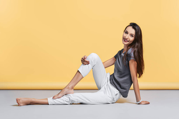Happy barefoot young woman in casual trousers and shirt sits on the floor in studio on yellow background - 写真・画像