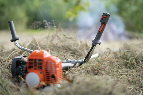 A handle with a start button for starting the engine of a petrol cutter close-up, blurred background. The petrol scythe trimmer brush cutter lies on the hay cut after mowing the grass in the garden. - Photo, Image
