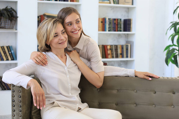 Beautiful middle aged mom and her adult daughter are hugging and smiling while sitting on couch at home - Valokuva, kuva