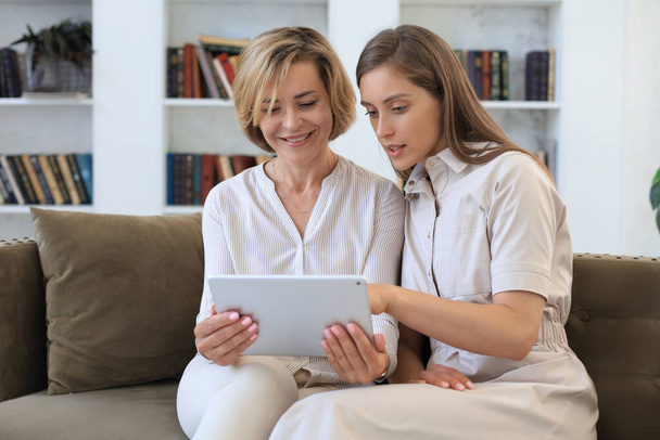 Happy mature woman with her adult daughter smiling while sitting on sofa and using digital tablet in living room - Photo, Image