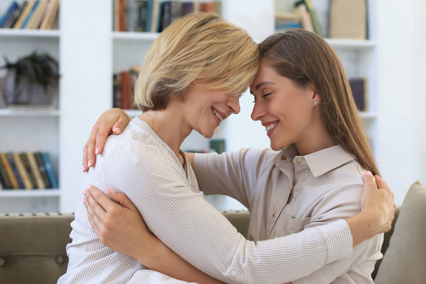 Cheerful young woman is embracing her middle aged mother in living room - Photo, image