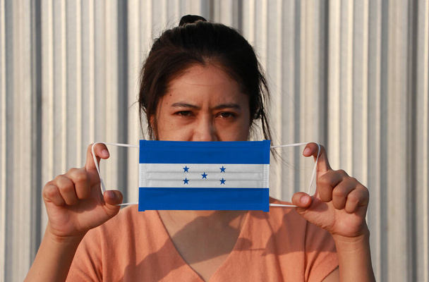 Egy nő Honduras zászlóval a kezében higiénikus maszkban, és felemelte a homlokát szürke háttérrel. Apró részecskék vagy víruskoronák vagy Covid 19 védelem. A betegség elleni küzdelem koncepciója. - Fotó, kép