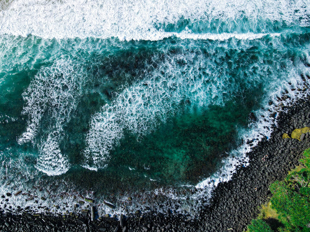 Hochauflösende Luftaufnahmen von Meereswellen an einer felsigen Küstenlinie mit grünen Regenwaldbäumen im Hintergrund. Kalter Winter blaugrünes Wasser und Steinstrand mit brechender Brandung - Foto, Bild