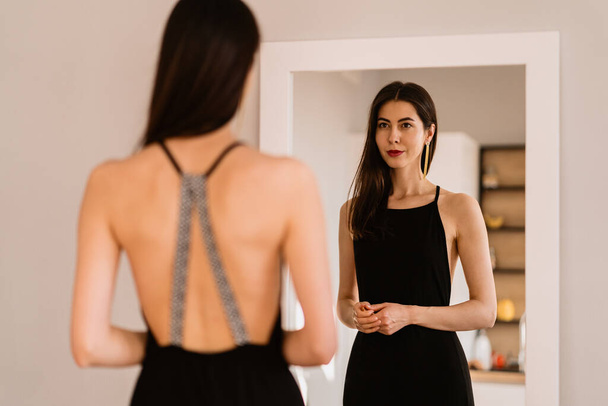 Η κυρία φοράει όμορφο μαύρο φόρεμα κοιτώντας στον καθρέφτη. - Φωτογραφία, εικόνα