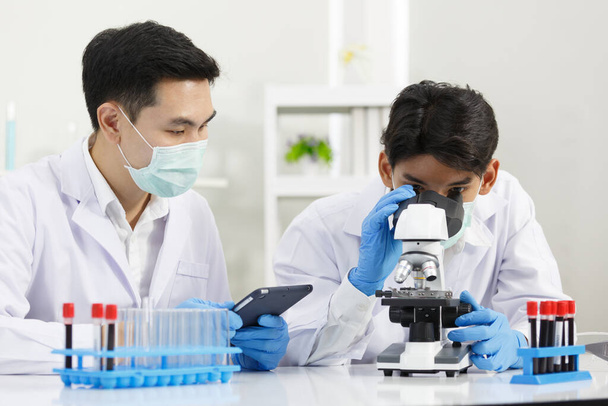Dva mužští vědci nosí masku obličeje pracující v laboratoři při kontrole řešení mikroskopem. SARS-CoV-2, Covid-19 THEME. - Fotografie, Obrázek