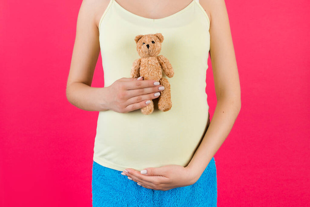 Primer plano de la mujer embarazada con ropa de hogar colorida sosteniendo un oso de peluche contra su vientre en el fondo rosa. La joven madre está esperando un bebé. Copiar espacio. - Foto, Imagen