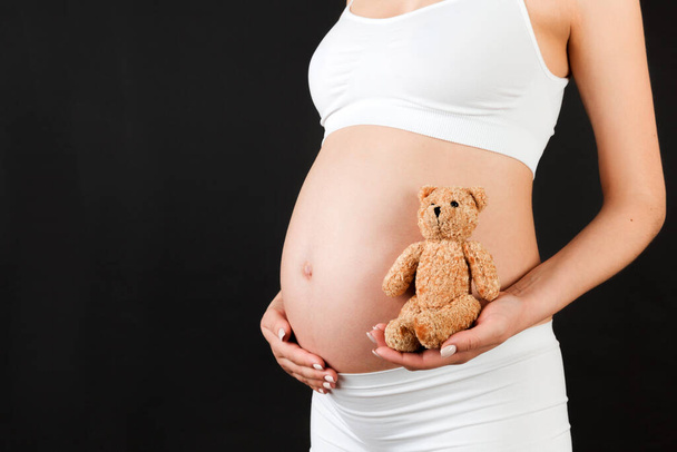 Close-up van teddybeer in de hand van zwangere vrouw op zwarte achtergrond. Toekomstige moeder draagt wit ondergoed. Naakte buik. Moederschap concept. Kopieerruimte. - Foto, afbeelding