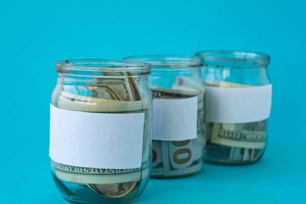 Three piggy bank from a glass jar with new dollars for life on a blue background. The banks have white sheets for inscriptions. Mockup. Copy space. Economy concept. - Foto, Bild