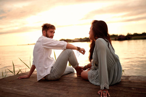 young beautiful caucasian couple sitting on wooden platform by the river, looking each other, talking - Foto, Imagem