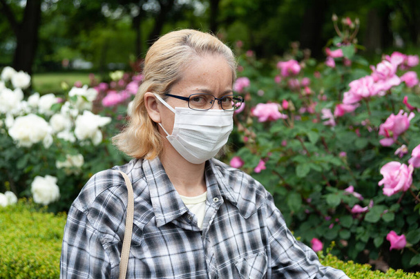 Retrato de mujer rubia mestiza de mediana edad con gafas con máscara quirúrgica blanca. Protección contra el coronavirus (COVID-19) y otras enfermedades infecciosas. Al aire libre. Mirada sospechosa. - Foto, Imagen