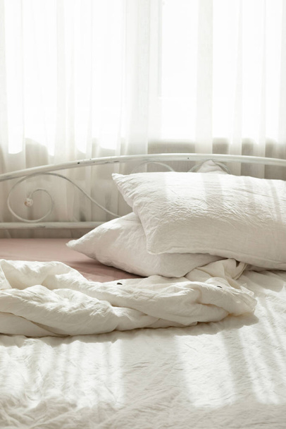 白いリネンのテキスタイルベッドウェア。枕の山。居心地の良いベッドルームのインテリアと美しい朝の光。パステルカラー。流行の有機天然リネン. - 写真・画像