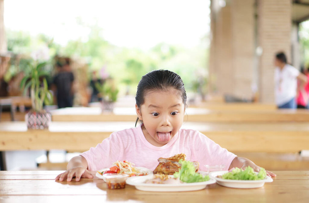 Enfant asiatique ou enfant fille wow et surprise stupéfaite avec profiter de manger du poulet grillé et salade de papaye pour la nourriture thaïlandaise et appétissant sur la table avec bâton langue et drôle de visage pour le déjeuner au restaurant - Photo, image