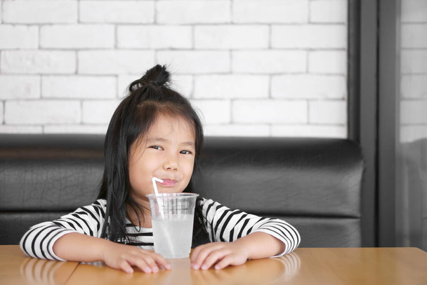 Asiatisches Kind niedlich oder kleines Mädchen lächelt und trinkt kühles und sauberes Wasser durch Schlauch oder Stroh im Plastikbecher für gesunde Frische mit Durst bei heißem Wetter im Sommer und sitzt auf dem Sofa zu Hause oder in der Schule und im Restaurant - Foto, Bild