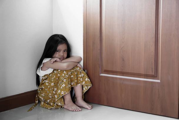 Aasian lapsi herkkä tai lapsi tyttö istuu halaus polvet ja kasvot kurtussa tai vihainen surullinen tai on ongelmia ja yksinäinen onneton tai oikukas etuovella ja huoneen nurkassa kotona tai päiväkodissa ja esikoulussa - Valokuva, kuva