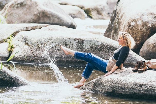 Γυναίκα με σακίδιο έχει ξεκουραστεί ενώ πεζοπορία στην κοιλάδα του ποταμού. Οικολογικός τουρισμός με σκανδιναβικούς πόλους. Νερό που πιτσιλάει με τα πόδια. - Φωτογραφία, εικόνα