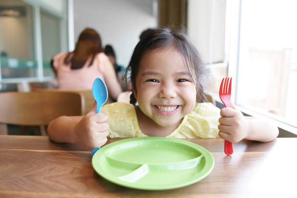 Asijské dítě roztomilé hladové nebo dítě dívka drží barevné lžíce a vidlička s jídlem na čekání jíst lahodné jídlo s úsměvem a šťastný pro vychutnat oběd nebo snídani v dopoledních hodinách v restauraci nebo jídelně - Fotografie, Obrázek