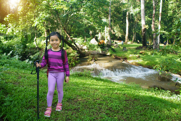 Asijské dítě nebo dítě dívka s úsměvem nést batoh nebo tašku a turistiku nebo vycházkové stožár pro trekking táboření stan na potoce se stromem v zelené trávě džungle pro dovolenou relaxovat a cestovat s dobrodružnou přírodou - Fotografie, Obrázek