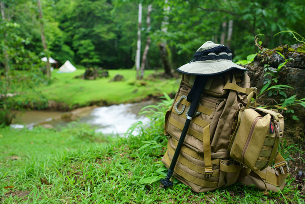 mochila con bolsa de medicina y senderismo o bastón con sombrero para trekking y tienda de campaña en arroyo con árbol en la selva de hierba verde para vacaciones relajarse y viaje de vacaciones con naturaleza aventura - Foto, Imagen