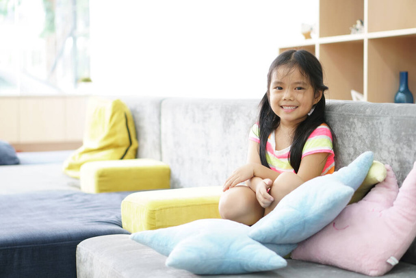 Aasian lapsi söpö tai lapsi tyttö hymyilee valkoiset hampaat ja onnellinen tai iloinen istua rentoutua sohvalla värikäs lepo tyyny kotona tai päiväkodissa ja päiväkodissa ajattelemalla uutta ideaa hammaslääkärin - Valokuva, kuva
